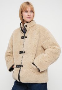 Veste beige en fausse peau retournée avec bordures noires, fermetures à boutons et grandes poches. Caractérisée par un col montant, une coupe décontractée et une texture duveteuse.