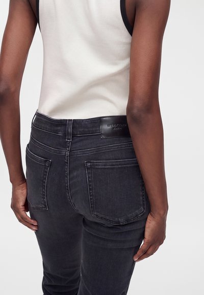 Personne portant un jean en denim noir et un haut blanc sans manches, vue de dos, avec des poches arrière visibles et un patch logo.