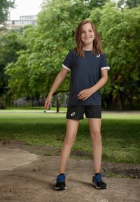 Laivastonsininen urheilupaita valkeilla yksityiskohdilla, mustat shortsit ja mustat urheilukengät sinisillä nauhoilla. Taustalla on vehreyttä ja betonipolkua.