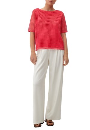 Femme portant un haut rouge en mesh à manches courtes, un pantalon blanc à jambes larges et des sandales marron à bout ouvert, debout devant un fond blanc.