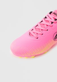 Φωτεινό ροζ ποδοσφαιρικό παπούτσι με κίτρινα πέλματα με καρφιά και κορδόνια, που διαθέτει μαύρο λογότυπο και διακριτικό κίτρινο σχέδιο στην περιοχή των δακτύλων.