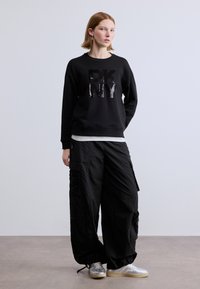 Μαύρο φούτερ με το λογότυπο "DKNY" από παγιέτες, συνδυασμένο με άνετο μαύρο παντελόνι cargo με τσέπες. Το μοντέλο φορά ασημένια αθλητικά παπούτσια.