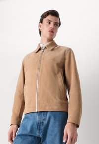 Tanfarbene Lederjacke mit glatter Textur, stehenden Kragen und Frontreißverschluss; kombiniert mit blauen Jeans.