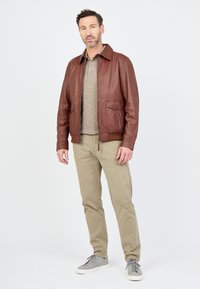 Braune Lederjacke mit Kragen, Fronttaschen und Reißverschluss. Trägt eine graue Hemd und ist mit beigen Hosen und grauen Turnschuhen kombiniert.