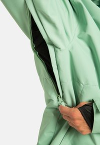 Ręka rozpinająca zamek wentylacyjny w jasnozielonej kurtce, pokazująca czarną siateczkową podszewkę wewnątrz.