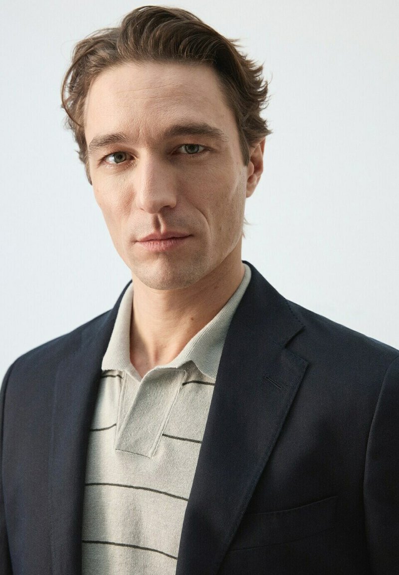 Homme aux cheveux bruns courts portant un blazer noir par-dessus un polo gris clair à fines rayures horizontales, expression neutre, fond blanc.