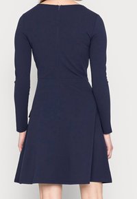 Marinblå långärmad klänning med figurnära liv och utsvängd kjol. Har en diskret dragkedja bak och ett mjukt, elastiskt material.