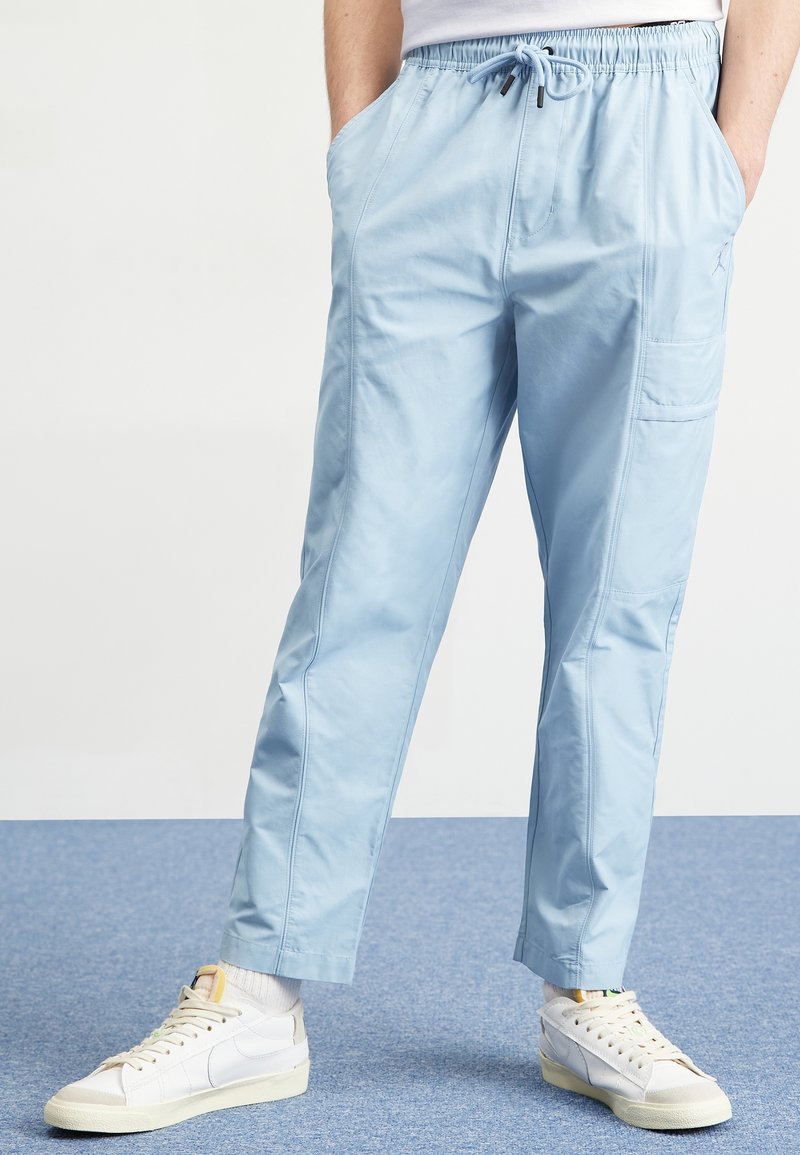 Personne portant un pantalon cargo bleu clair avec une taille élastique à cordon et des baskets blanches, debout sur un tapis bleu.