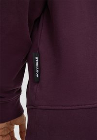 Violetter Sweatshirt aus strukturiertem Stoff, Nahaufnahme des Bündchens und Saums, mit einem schwarzen Etikett, das die Marke "Strellson" zeigt.