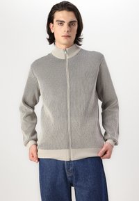 Lys grå ribbet zip-up sweater med høj krave og lange ærmer, parret med blå denim jeans. Struktureret stof med en tætsiddende design.