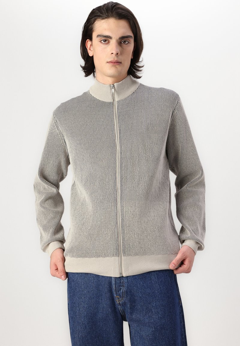 Maglione grigio chiaro a coste con zip, colletto alto e maniche lunghe, abbinato a jeans in denim blu. Tessuto testurizzato con un design aderente.