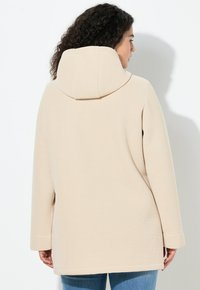 Béžová mikina s kapucí z texturované látky, dlouhými rukávy a volným střihem. Má velkou, pevnou kapuci a boční rozparky na lemu.