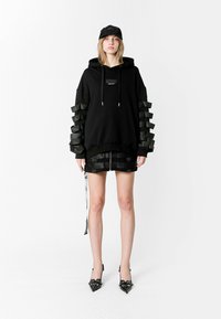 Sudadera negra con grandes acentos de goma en las mangas, combinada con una minifalda plisada negra y tacones de punta. La gorra negra completa el look.