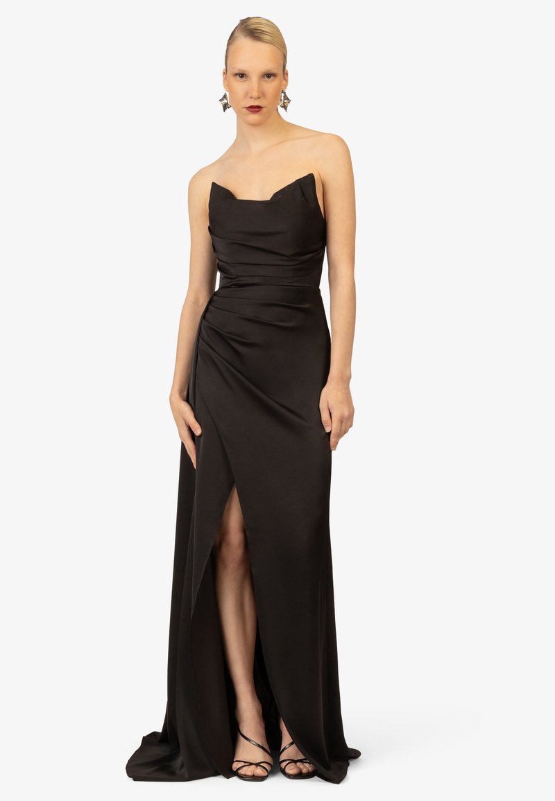 Schwarzes Satin-Abendkleid mit Herzausschnitt, gerafften Details und einem hohen Schlitz, kombiniert mit schwarzen Sandalen mit Riemen.