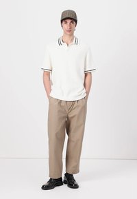 Biely textured polo tričko s čiernymi pruhmi na golieri, skombinované s béžovými voľnými nohavicami. Model nosí károvanú čiapku a čierne topánky.