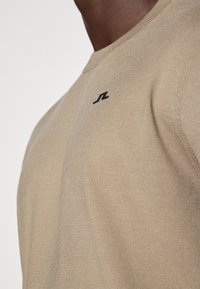 Bežna pletena majica s crewneck izrezom z mehko teksturo, ki ima majhen črn logotip na prsih. Poudarjen pogled na podrobnosti tkanine.
