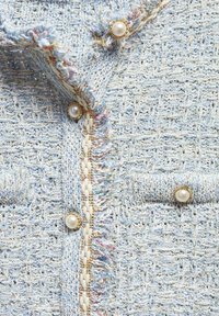 Tissu bleu clair texturé avec un motif tissé, comprenant des boutons ornés de perles et une bordure à franges le long des bords.