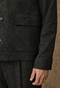 Sacou cu model în zig-zag negru, dotat cu un buzunar și butoane, realizat dintr-o țesătură texturată. Pantalonii asortați sunt vizibili în partea de jos.