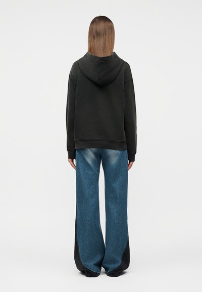 Άτομο με ίσια καστανά μαλλιά, ντυμένο με μαύρη φούτερ με κουκούλα και φαρδιά μπλε τζιν, εμφανίζεται από πίσω με φόντο ένα απλό υπόβαθρο.