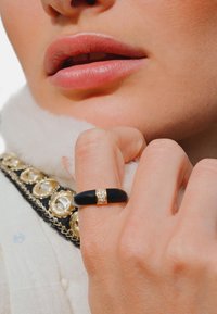 Bague en céramique noire avec un accent doré comprenant de petites cristaux incrustés. Portée sur une main à la peau claire, aux côtés d'un col blanc texturé.