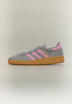 Γκρι αθλητικό Adidas με ροζ ρίγες και φτέρνα, χρυσή ετικέτα Spezial, καφέ σόλα και λευκά κορδόνια σε απλό ανοιχτό φόντο.