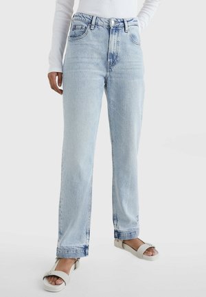 Personne portant un jean droit taille haute bleu clair, un haut blanc à manches longues et des sandales blanches ouvertes à l'avant avec une bride à boucle.