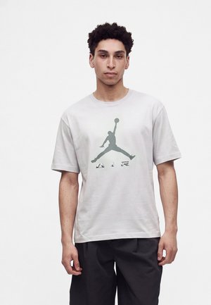 Mladý muž s krátkymi kučeravými vlasmi, ktorý má na sebe biele tričko s veľkým sivým logom Jumpman a čierne nohavice, stojí pred jednoduchým bielym pozadím.