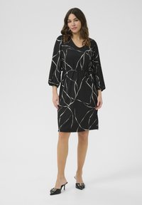 Robe noire avec des lignes abstraites blanches, décolleté en V, taille élastique et manches ballons trois quarts. Portée avec des mules à talons noirs.