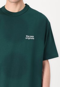 Mørkegrønn bomulls t-skjorte med korte ermer og rund hals. Har hvit brodert tekst "de nye originalene" på venstre bryst.