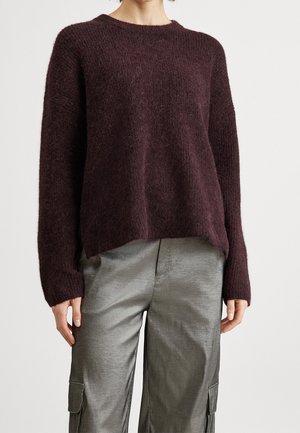 Person iført en mørkelilla, blød langærmet sweater og grå cargobukser med lommer, stående foran en ensfarvet hvid baggrund.