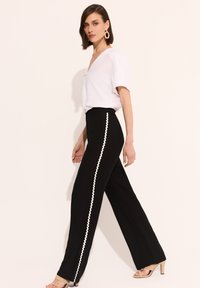 Czarne spodnie z szerokimi nogawkami z białą, falbanistą lamówką po bokach, w połączeniu z białą bluzką z krótkim rękawem i beżowymi sandałami na wysokim obcasie.