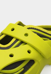 Κοντινό πλάνο ενός φωτεινού κίτρινου λαστιχένιου σανδάλου με ρυθμιζόμενο λουράκι και οπές αερισμού, που αποκαλύπτει υφή μαύρου υφάσματος από κάτω.