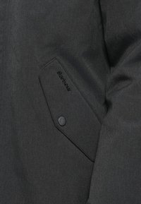 Μαύρο εξωτερικό μπουφάν με υφή υφάσματος, διαγώνια τσέπη με κουμπί snap και ανάγλυφο λογότυπο της μάρκας Barbour στην τσέπη.