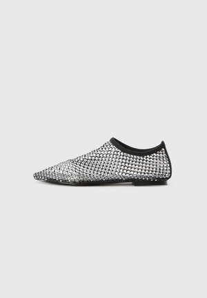 Chaussure slip-on noire et argentée avec un bout pointu, présentant un motif en maille et une cheville élastiquée. Semelle plate avec peu de matériel.