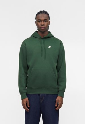 Junger Mann mit geflochtenem Haar, der einen dunkelgrünen Nike-Hoodie und dunkelblaue Jeans trägt, steht mit Blick nach vorne vor einem weißen Hintergrund.