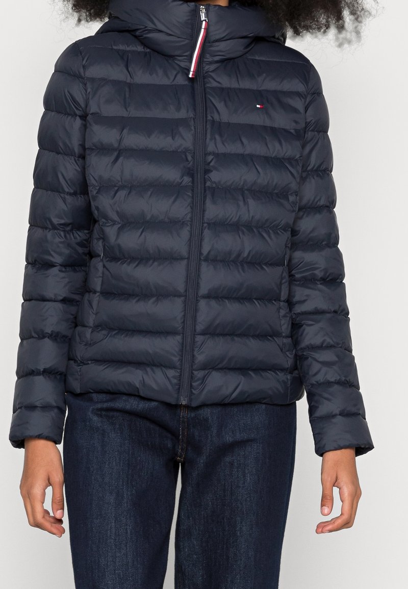 Veste rembourrée marine avec un design matelassé, capuche et fermeture éclair à l'avant. Elle dispose de poignets élastiques et d'un détail logo sur la poitrine.