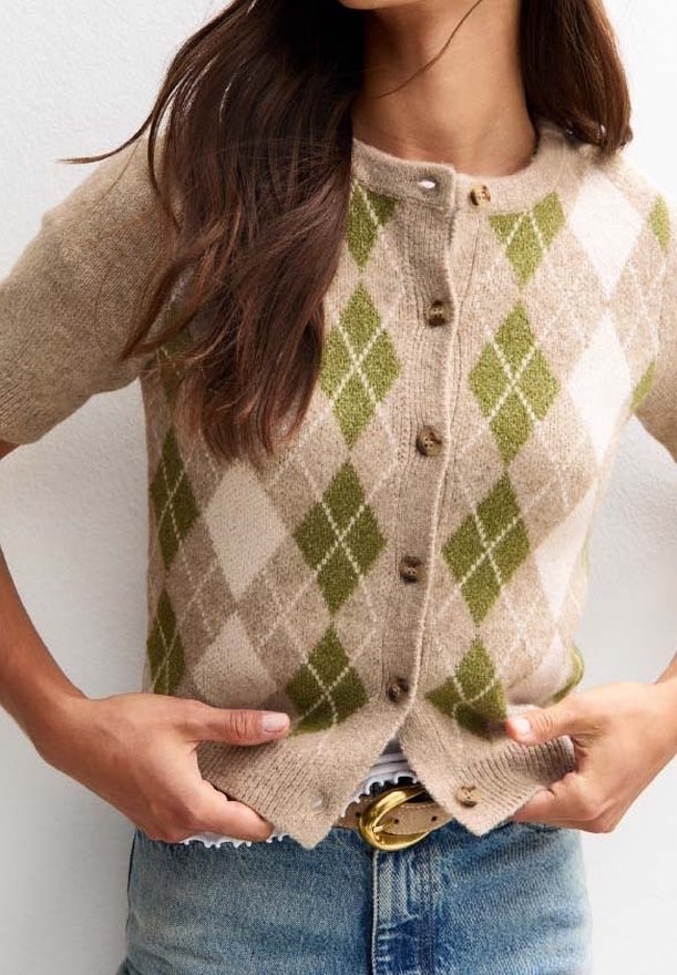 Femme portant un cardigan beige à manches courtes avec un motif losanges vert et blanc, boutonné devant, associé à un jean bleu et une ceinture.