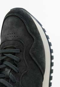 Sneaker nera in suede con tomaia texturizzata, punta arrotondata, lacci piatti e suola in gomma con motivo. Presenta una suola intermedia bianca a contrasto.