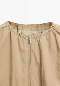 Beige lightweight jacket with a silver front zipper and gathered elastic neckline on a white background.