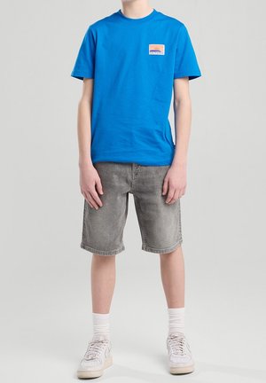 Person trägt ein hellblaues T-Shirt mit einem California-Aufnäher, graue Jeansshorts, weiße Socken und weiße Turnschuhe und steht vor einem einfarbigen Hintergrund.