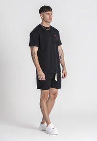 Černé tričko s krátkým rukávem a malým červeným logem, spárované s černými šortkami a bílými teniskami. Má volný střih a šňůrkový pas.