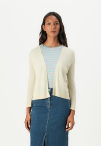 Cardigan beige clair avec un col châle, des manches longues et des bords côtelés, superposé à un haut rayé bleu et blanc. Jupe en denim avec une fente à l'avant.