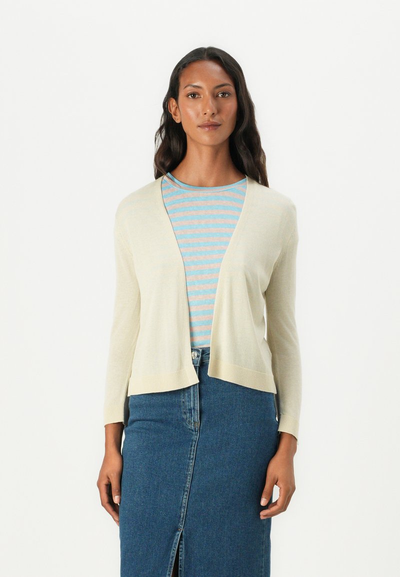 Cardigan beige clair avec un col châle, des manches longues et des bords côtelés, superposé à un haut rayé bleu et blanc. Jupe en denim avec une fente à l'avant.