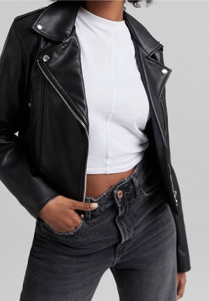 Person iført en sort læderjakke over en hvid croppet top med hånden i lommen på højtaljede, falmede sorte jeans, grå baggrund.