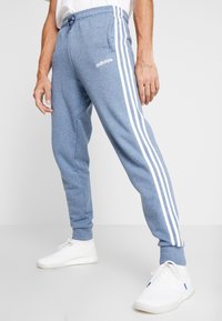Modré jogger kalhoty ze směsi bavlny s bílými bočními pruhy a elastickým pasem. Na levém stehně je logo adidas. Stylizováno s bílými teniskami.