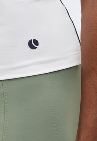 Λευκό αθλητικό πουκάμισο με υφή και μαύρο λογότυπο σε σχήμα ημισελήνου πάνω από πράσινο μέντας κολάν στο σώμα και το χέρι ενός ατόμου.