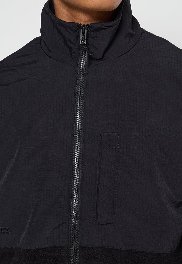 Veste zippée noire avec une surface texturée, col montant et poche avant. Équipée d'une fermeture éclair lisse et d'un tissu inférieur contrastant.