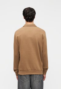 Καφέ πουλόβερ με γιακά, φτιαγμένο από μαλακό ύφασμα. Διαθέτει μακριά μανίκια και εφαρμοστή γραμμή, με ριμπ μανσέτες και τελείωμα. Μεγάλες, ανοιχτό γκρι πίσω τσέπες ορατές.