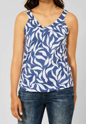 Vrouw draagt een blauw mouwloos topje met wit bladmotief en donkerblauwe jeans, staand tegen een effen achtergrond.