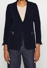 Blazer bleu marine ajusté en tissu léger, doté d'un bouton unique, de deux poches frontales zippées et d'un col cranté.